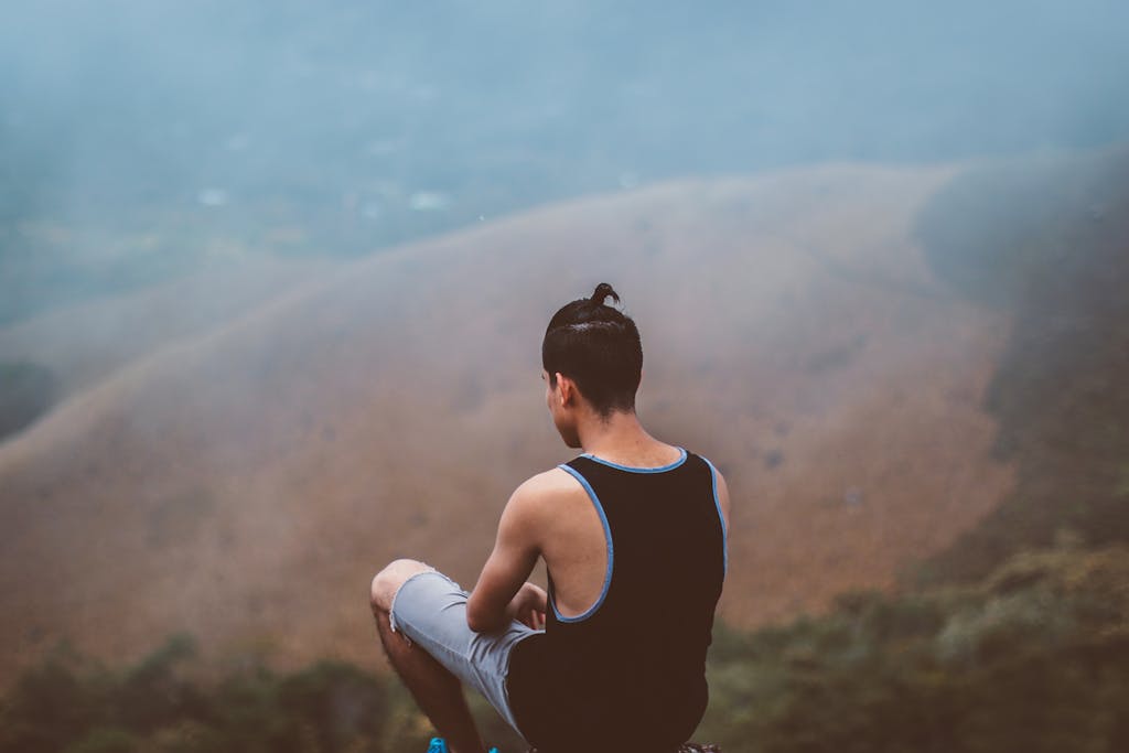 A man sits on the edge of a foggy mountain, surrounded by nature in a tranquil setting.